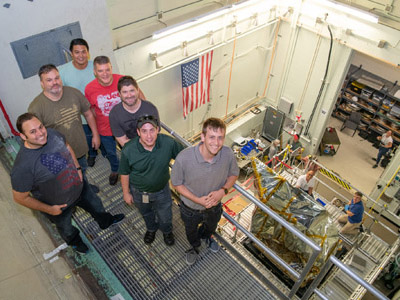 The Ocean Color Instrument mechanical team stands on the balcony overlooking the bagged instrument after successfully installing it onto the X axis (vertical) vibration shaker table. Credit: Stover, Desiree